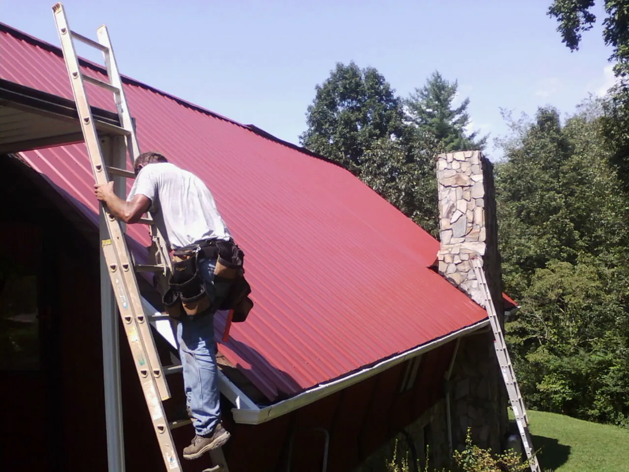 Roofer on a ladder beside a red metal roof and stone chimney — Soffit & Fascia Repair by Roofing Brothers in Caruthers